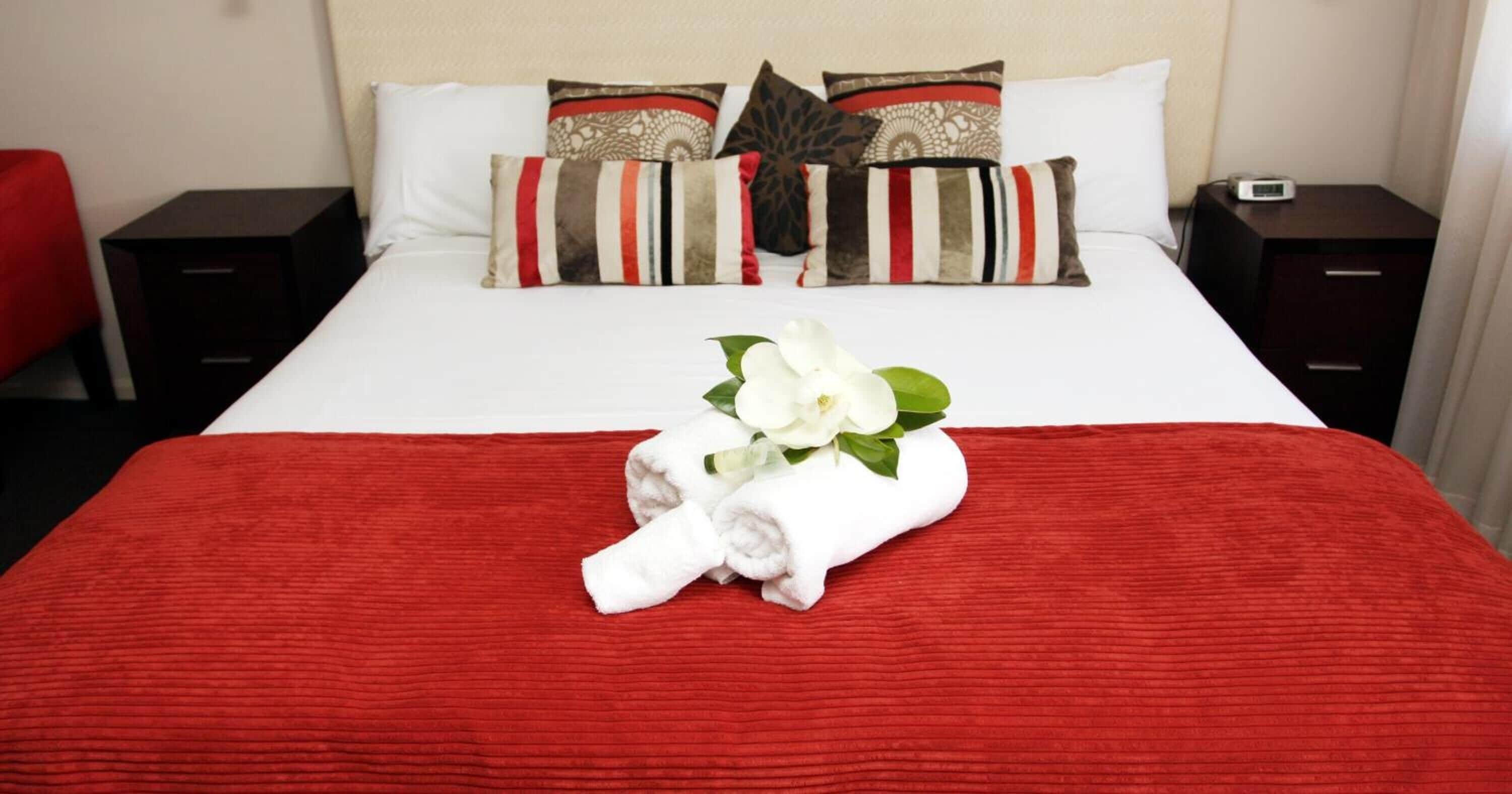 A neatly made bed with a red blanket, white pillows, and a bouquet of white flowers.