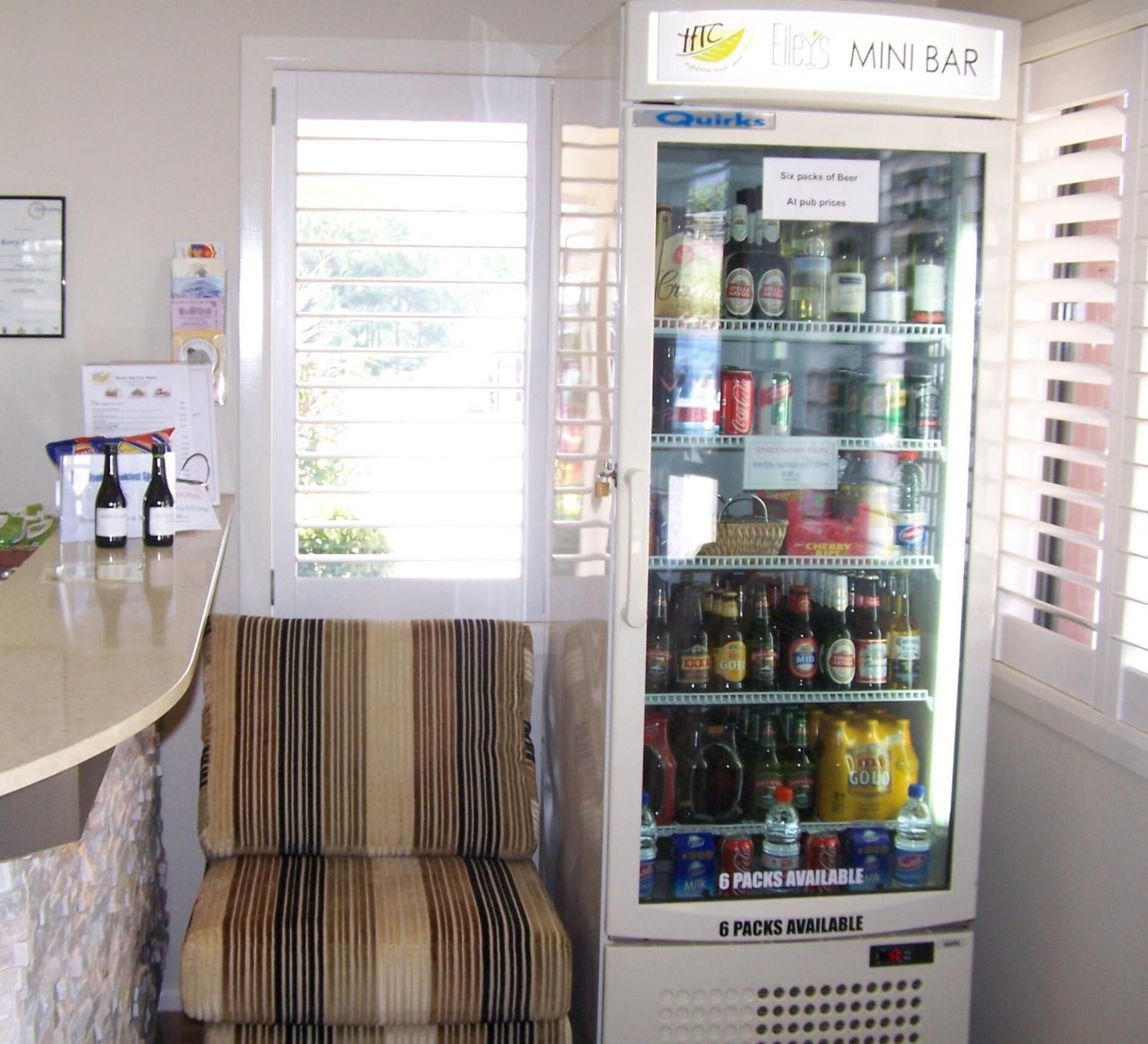 Un minibar con una variedad de bebidas, incluyendo cerveza, refrescos y agua. Hay una silla a rayas frente al minibar.