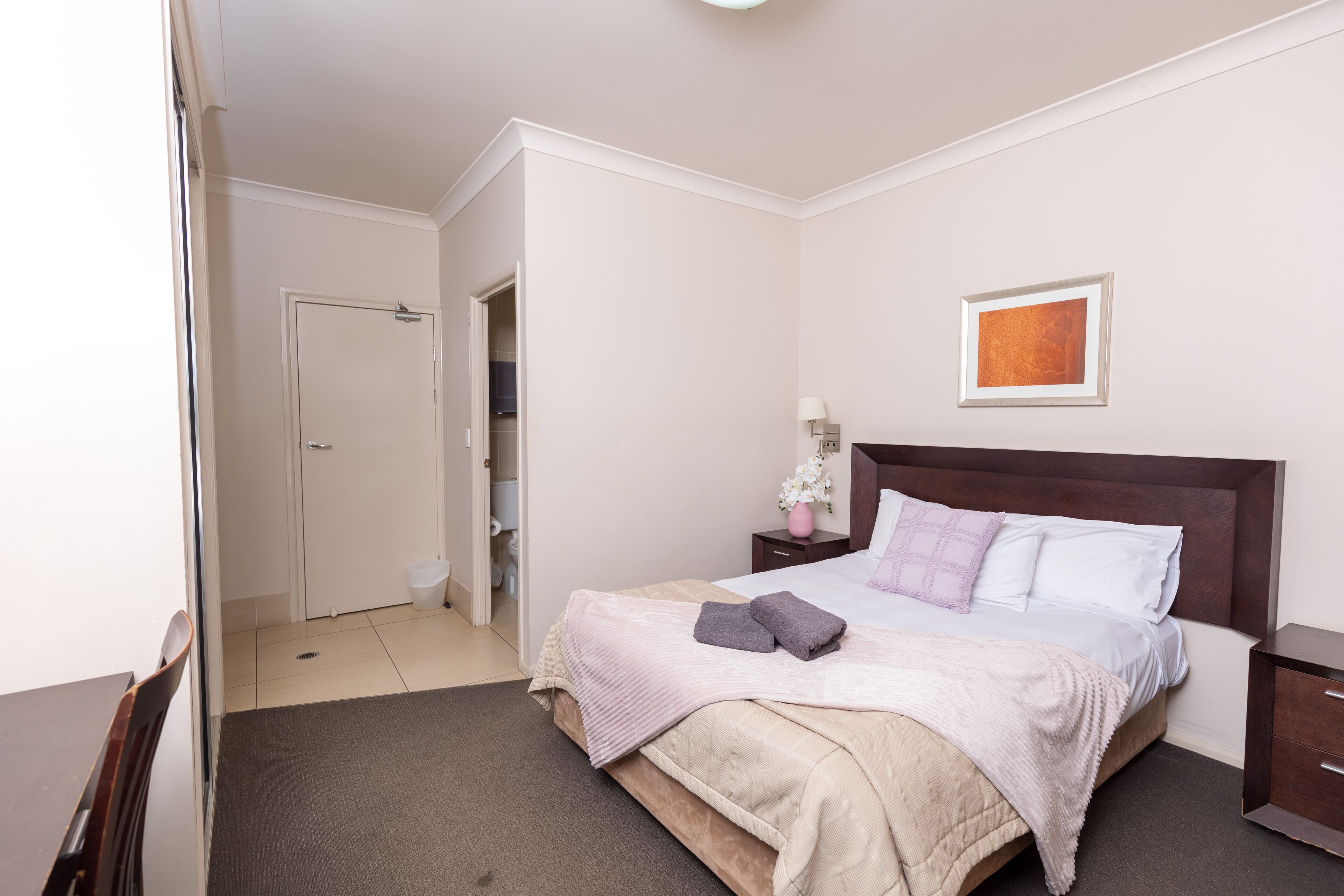 A neatly arranged bedroom with a bed, bedside tables, a nightstand, a framed picture on the wall, and a door leading to another room.