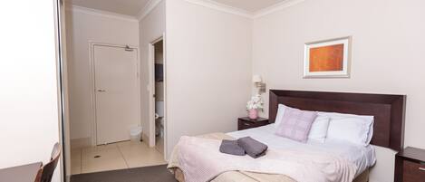 A neatly arranged bedroom with a bed, bedside tables, a nightstand, a framed picture on the wall, and a door leading to another room.