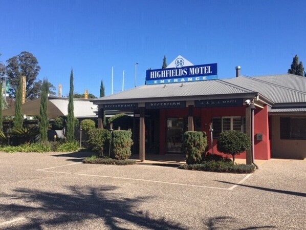 The Highfields Motel entrance with a clear sky, a parking lot, and a building with a red facade.