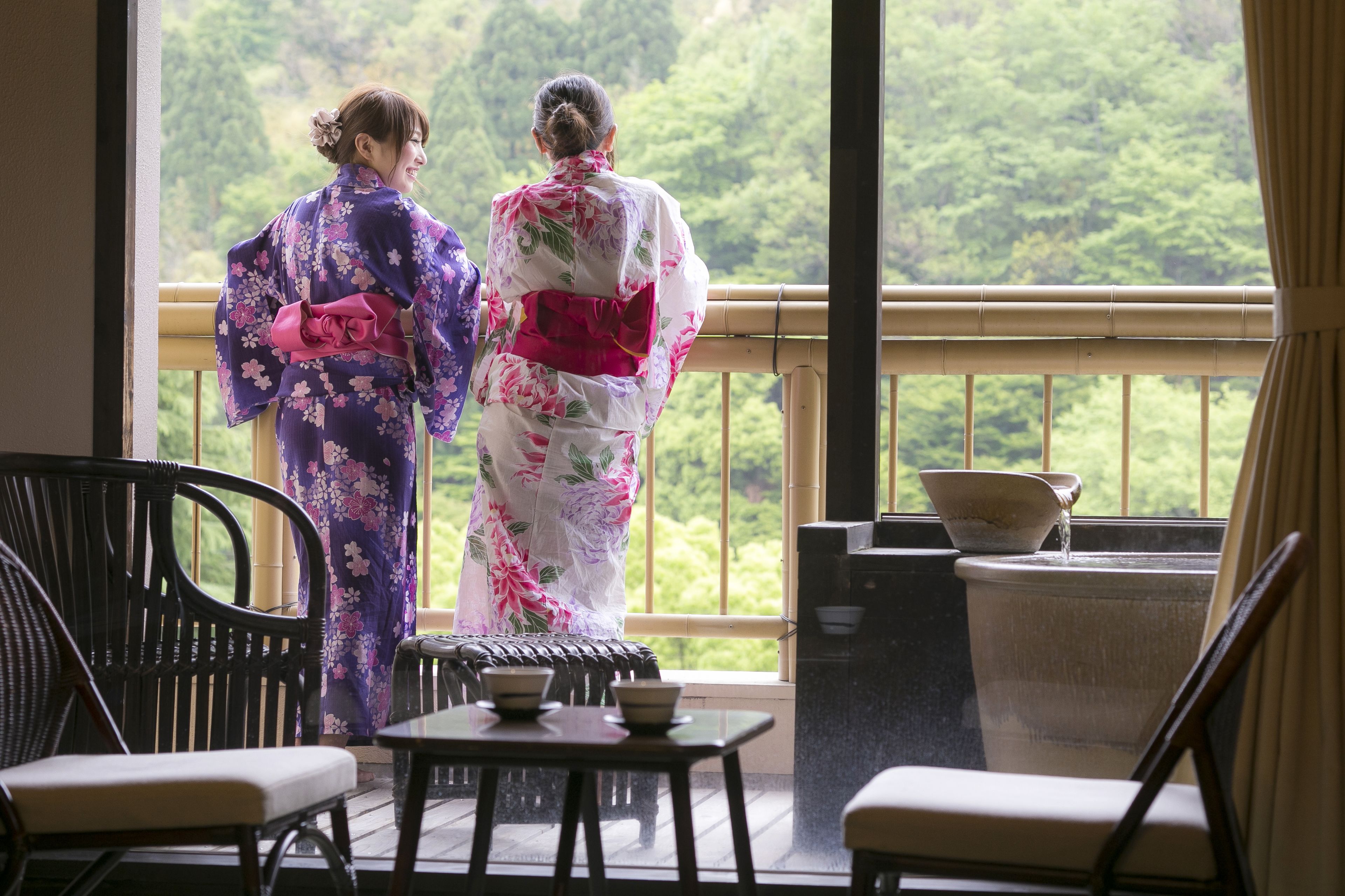 A traditional Japanese room with tatami flooring, a low table with a tea set, and a view of lush greenery through large windows.