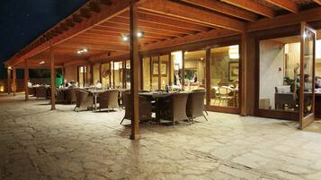 An outdoor covered patio with stone flooring and wooden beams, featuring tables and chairs.