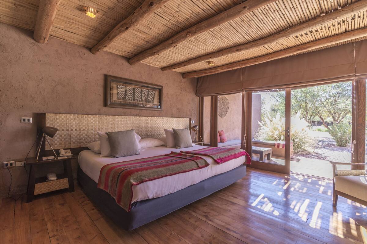 A spacious bedroom with a large bed, wooden flooring, a ceiling with exposed beams, and a view of the outdoors through a sliding glass door.