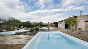 A pool area with wooden decking and a clear blue pool, surrounded by greenery and a building with a rustic facade.