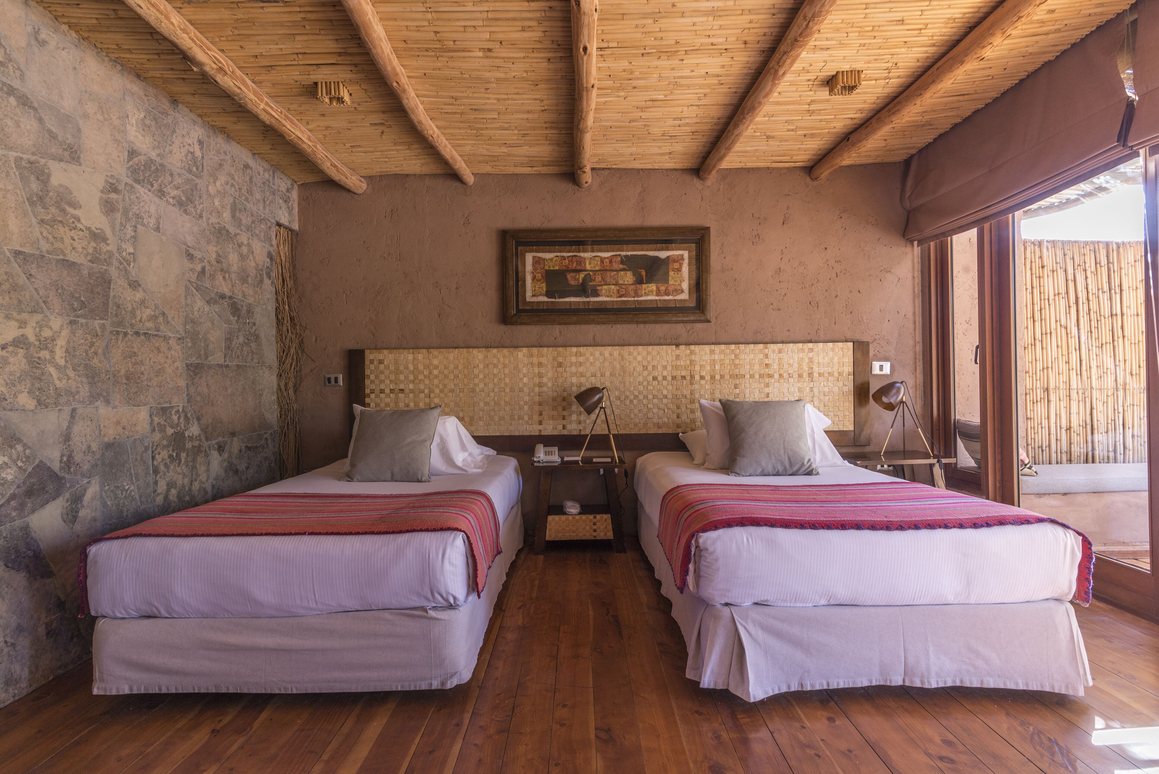 A room with two beds, wooden flooring, a stone wall, and a large window.