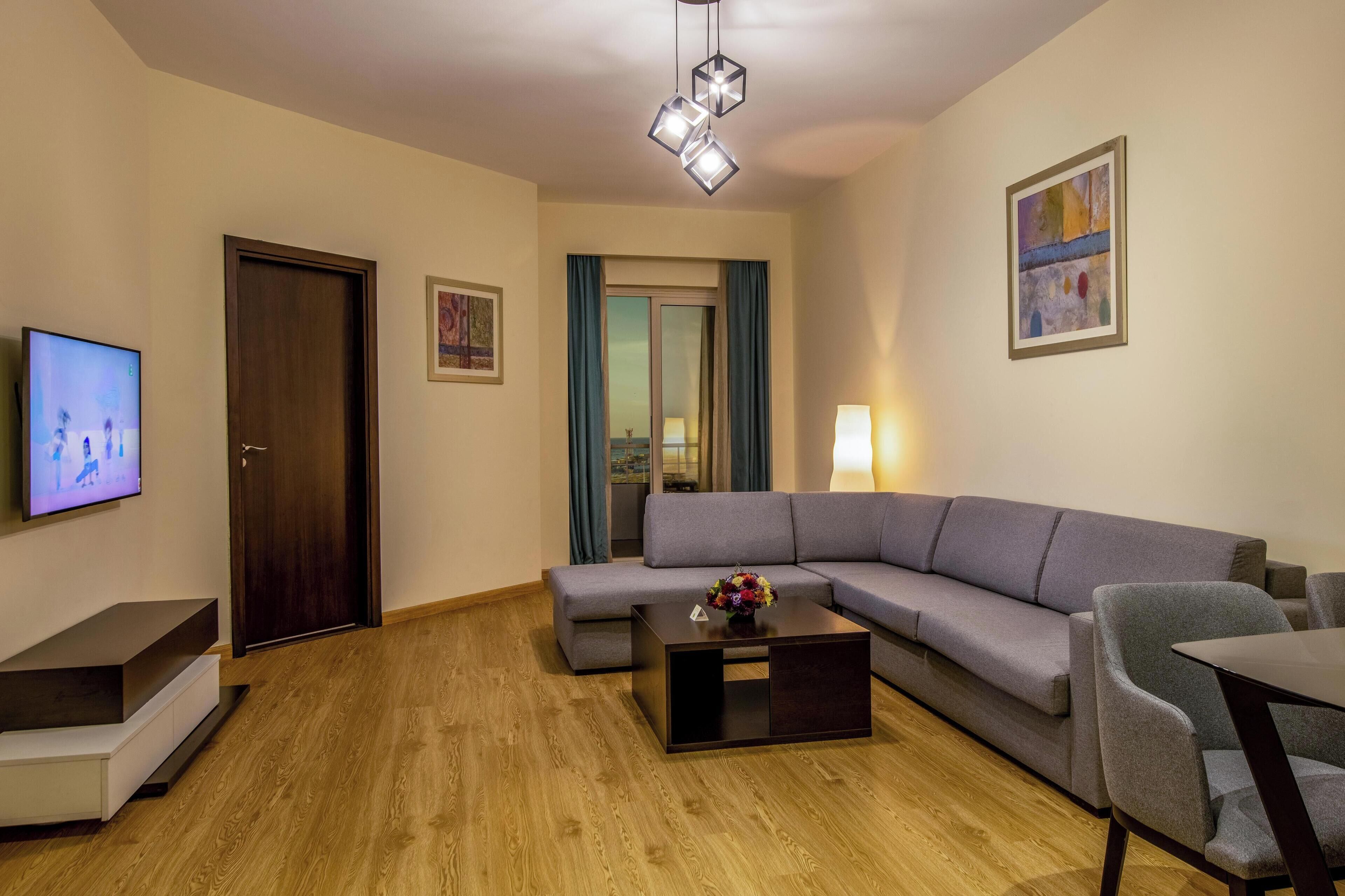 Un salon moderne avec un canapé gris, une table basse ornée d’un vase de fleurs, un téléviseur fixé au mur et un plancher en bois.