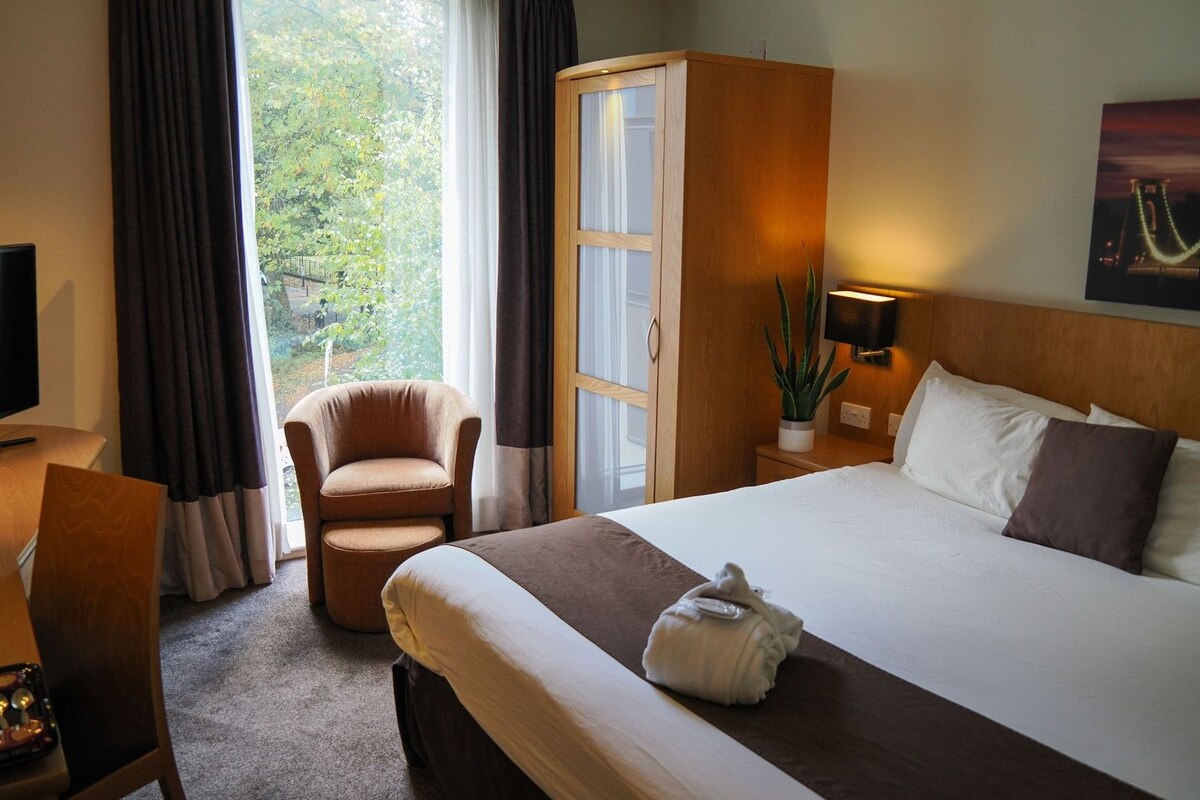 Una habitación de hotel con cama, una silla, un escritorio con computadora, un armario y una ventana con cortinas.