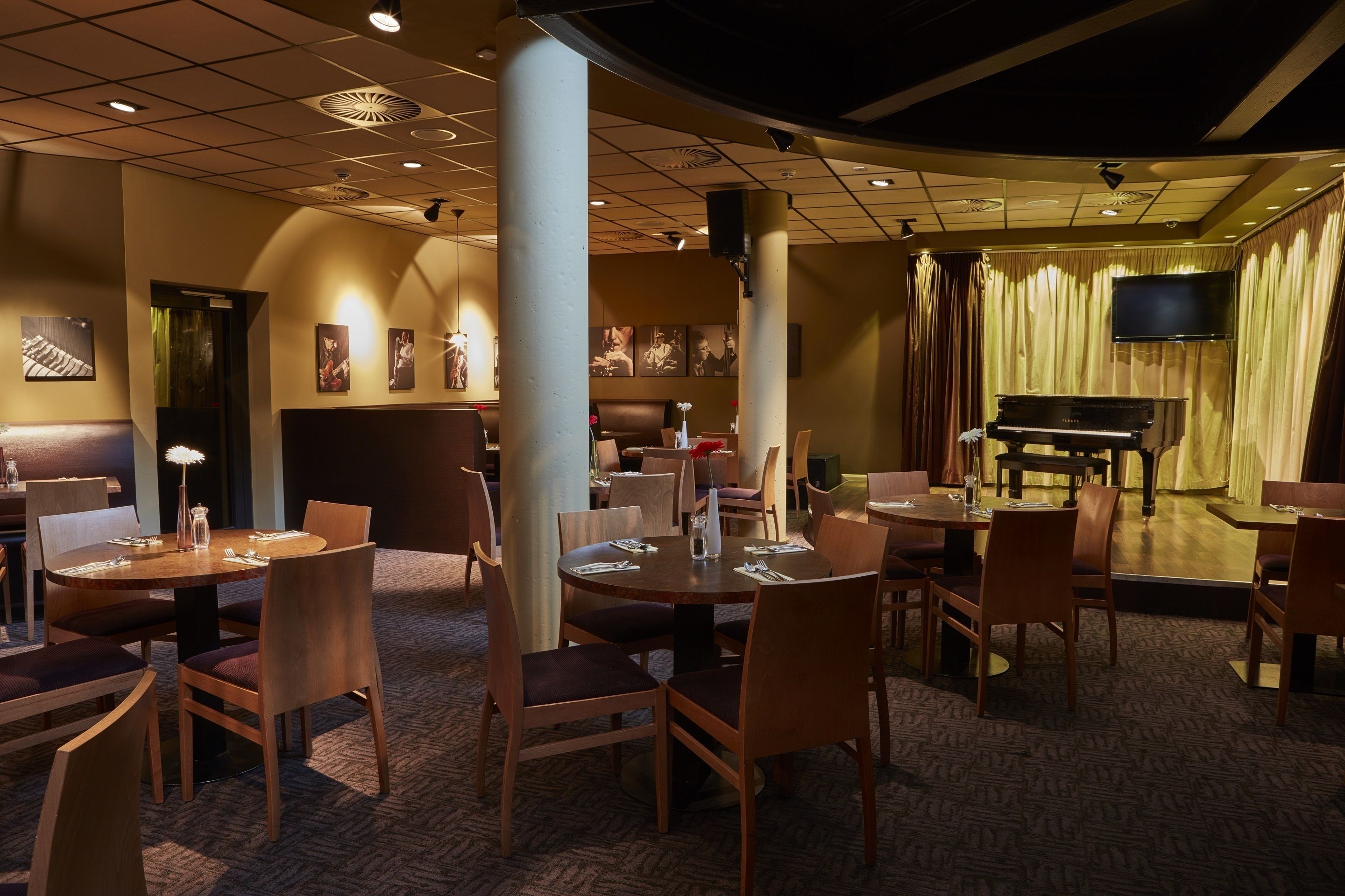 A restaurant with round tables and chairs, a piano, and a TV on the wall.