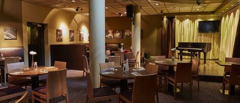 A restaurant with round tables and chairs, a piano, and a TV on the wall.
