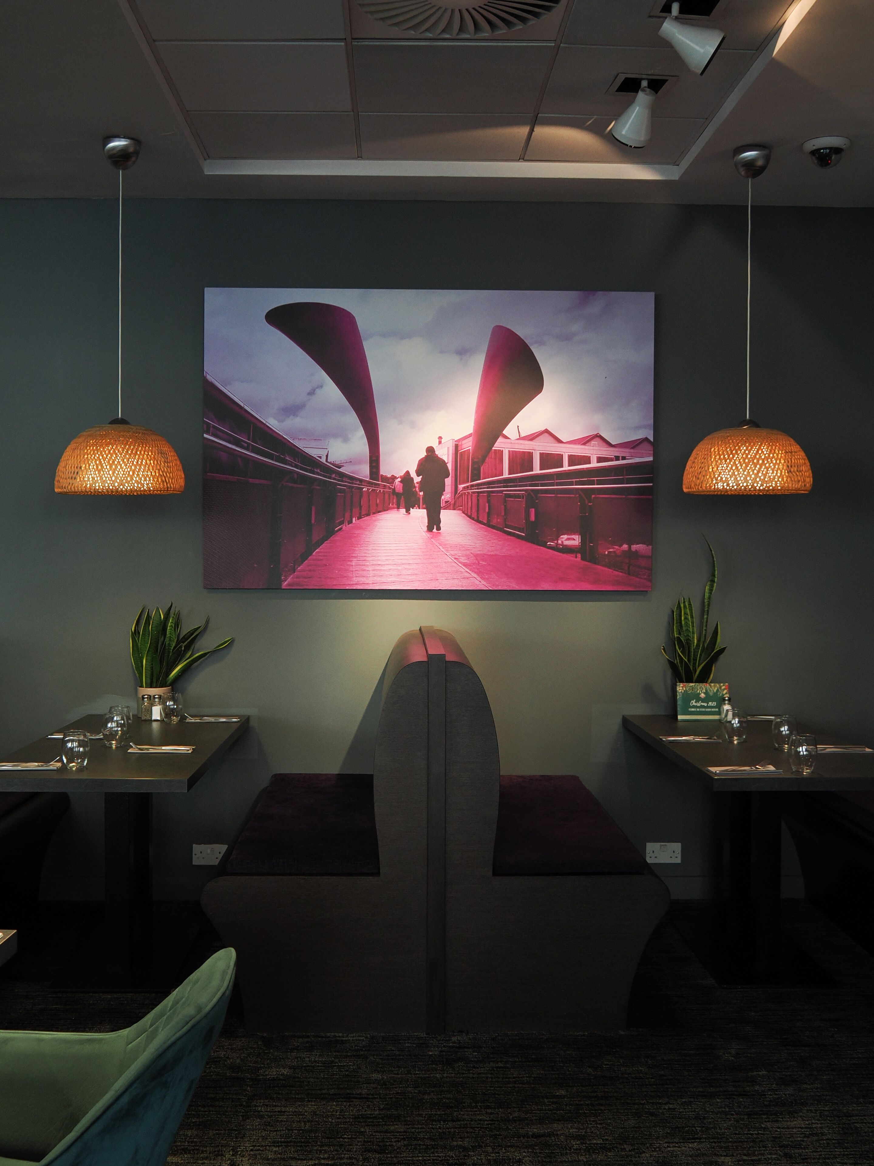 Un intérieur moderne agrémenté d’une grande œuvre d’art murale représentant un pont, de deux tables à manger ornées de plantes en pot et d’un éclairage suspendu.