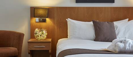 A hotel room with a wooden headboard, a bedside lamp, a vase of flowers, a neatly made bed with white linens, and a framed picture on the wall.