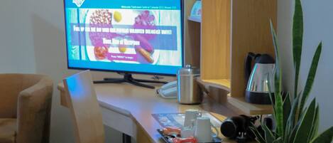 A hotel room with a wooden desk, a computer monitor, a coffee maker, a plant, and a chair.