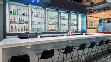 Zona de bar con un letrero de neón, varias estanterías llenas de botellas y taburetes negros.