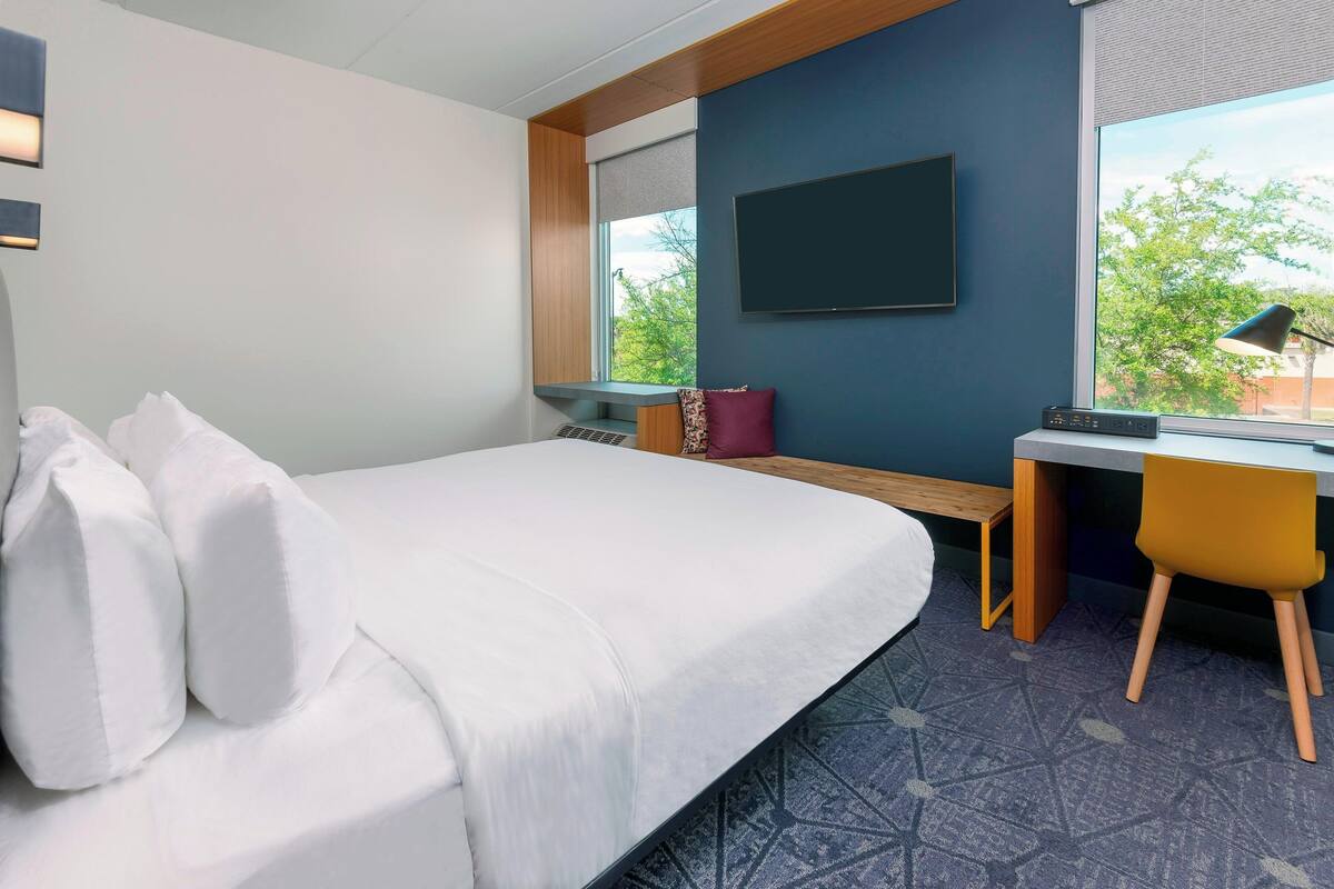 Une chambre d’hôtel avec un lit, un bureau, une chaise, une télévision et une fenêtre donnant sur des arbres.