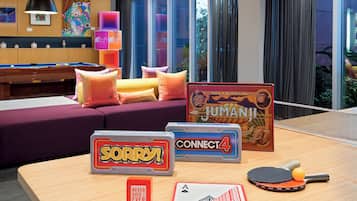 Una mesa de billar junto a una mesa de ping pong, un juego de cartas y un sofá en una habitación con ventanales amplios.