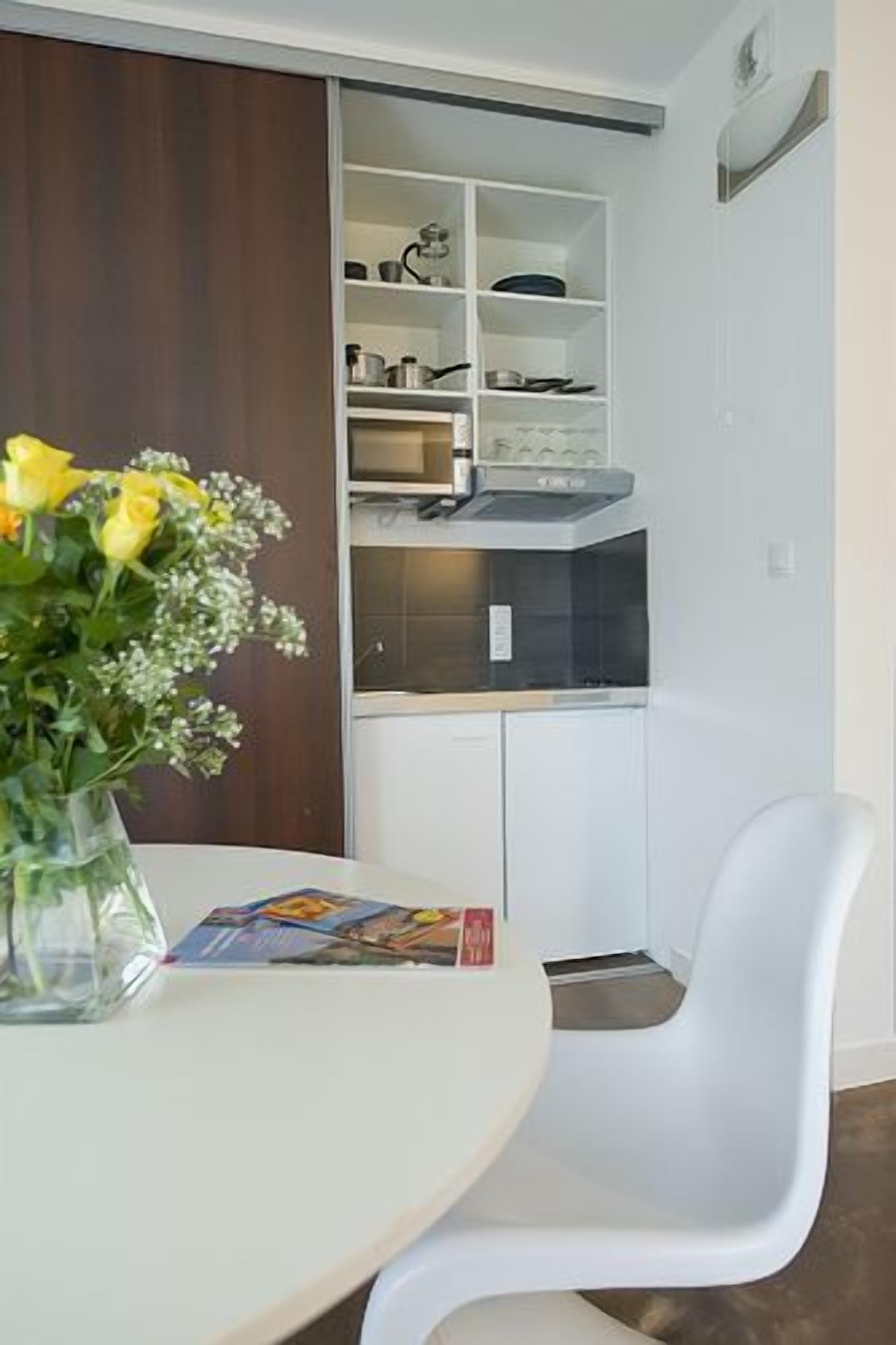 A compact kitchen with white cabinets, a microwave, and a sink. There's a white chair and a table with a vase of yellow flowers.