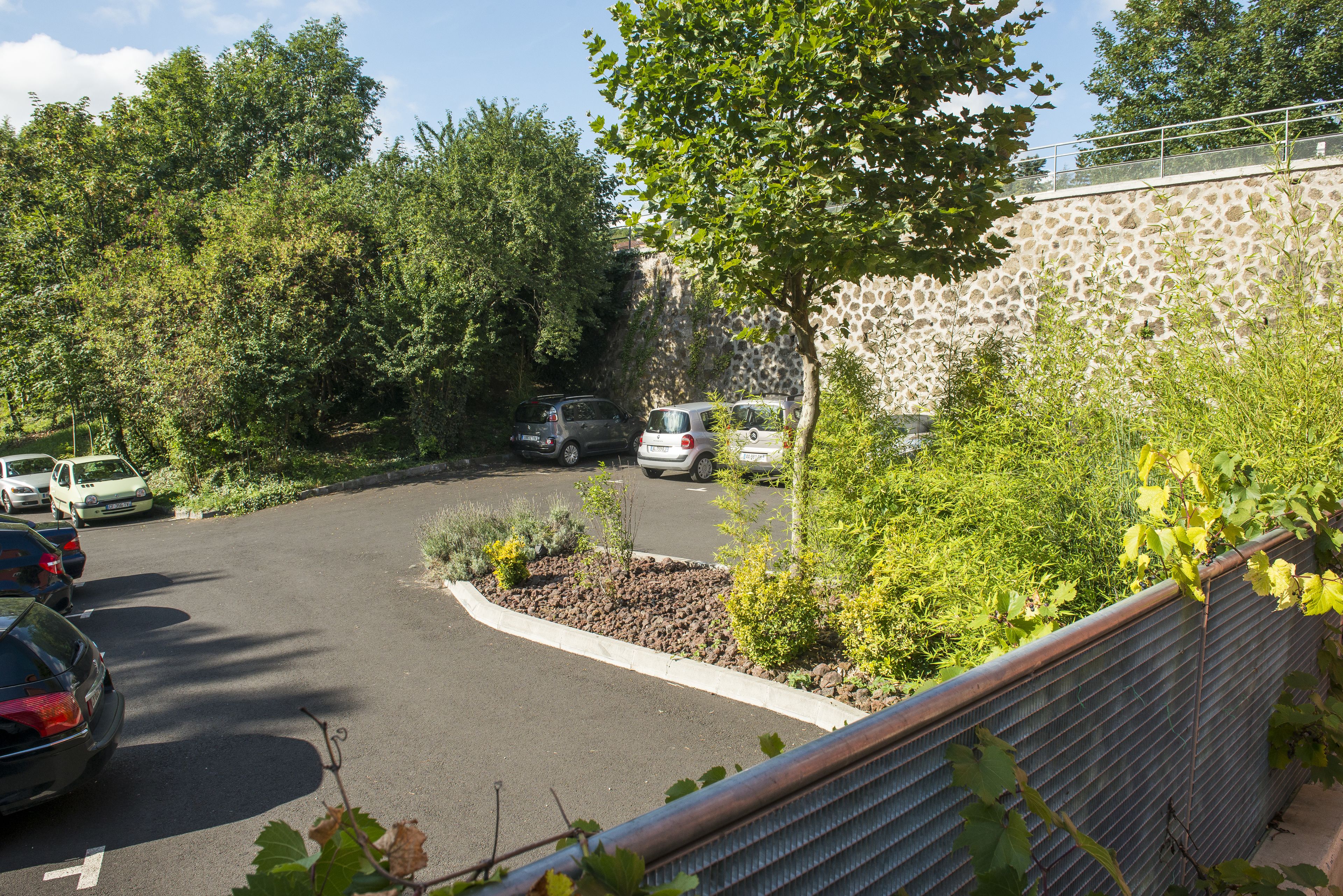 A parking area with cars, surrounded by greenery and a stone wall.