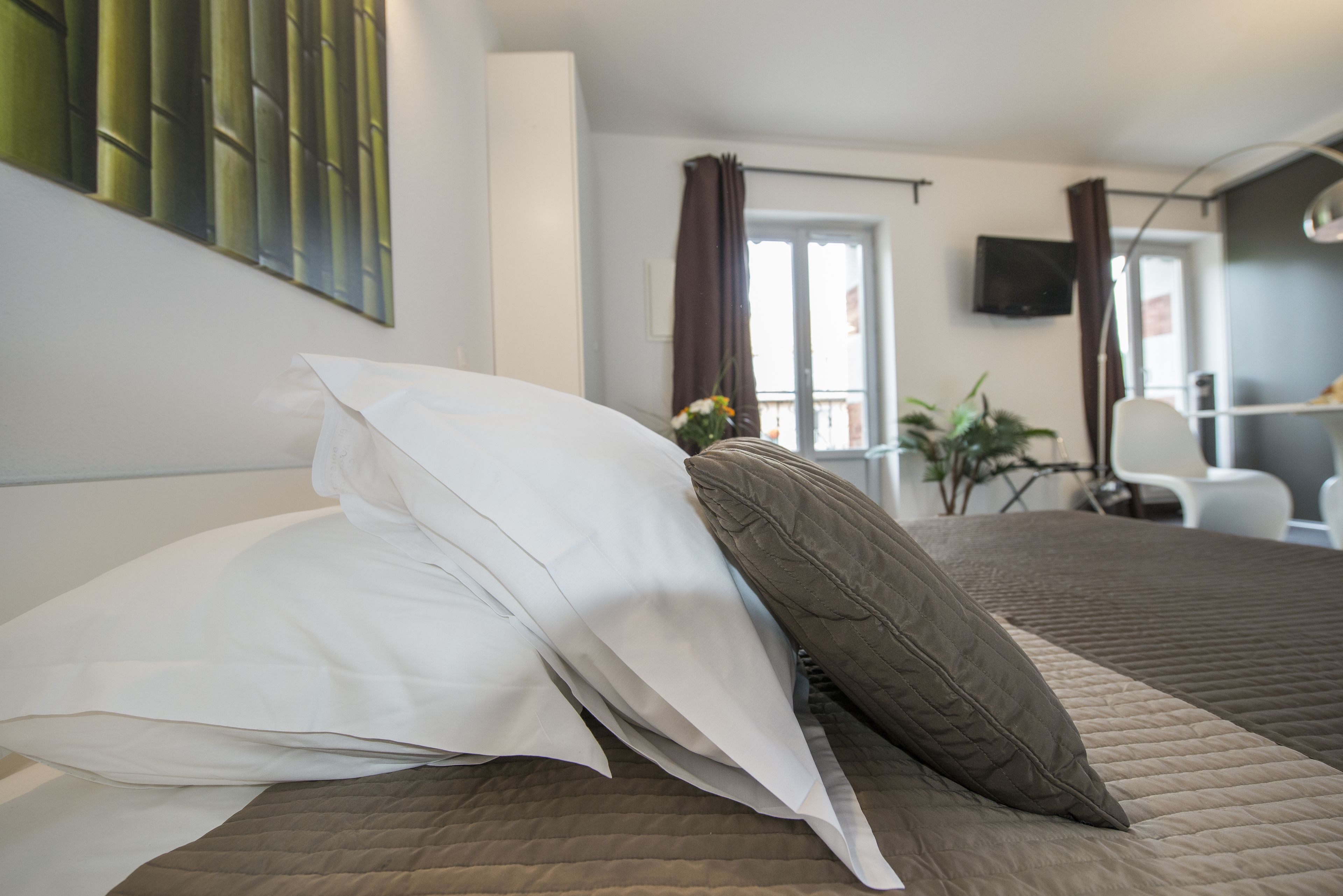 A neatly made bed with white and brown pillows, a quilted brown bedspread, and a view of a balcony with a plant and a chair.