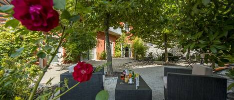 An outdoor seating area with tables and chairs, surrounded by greenery and a building in the background.