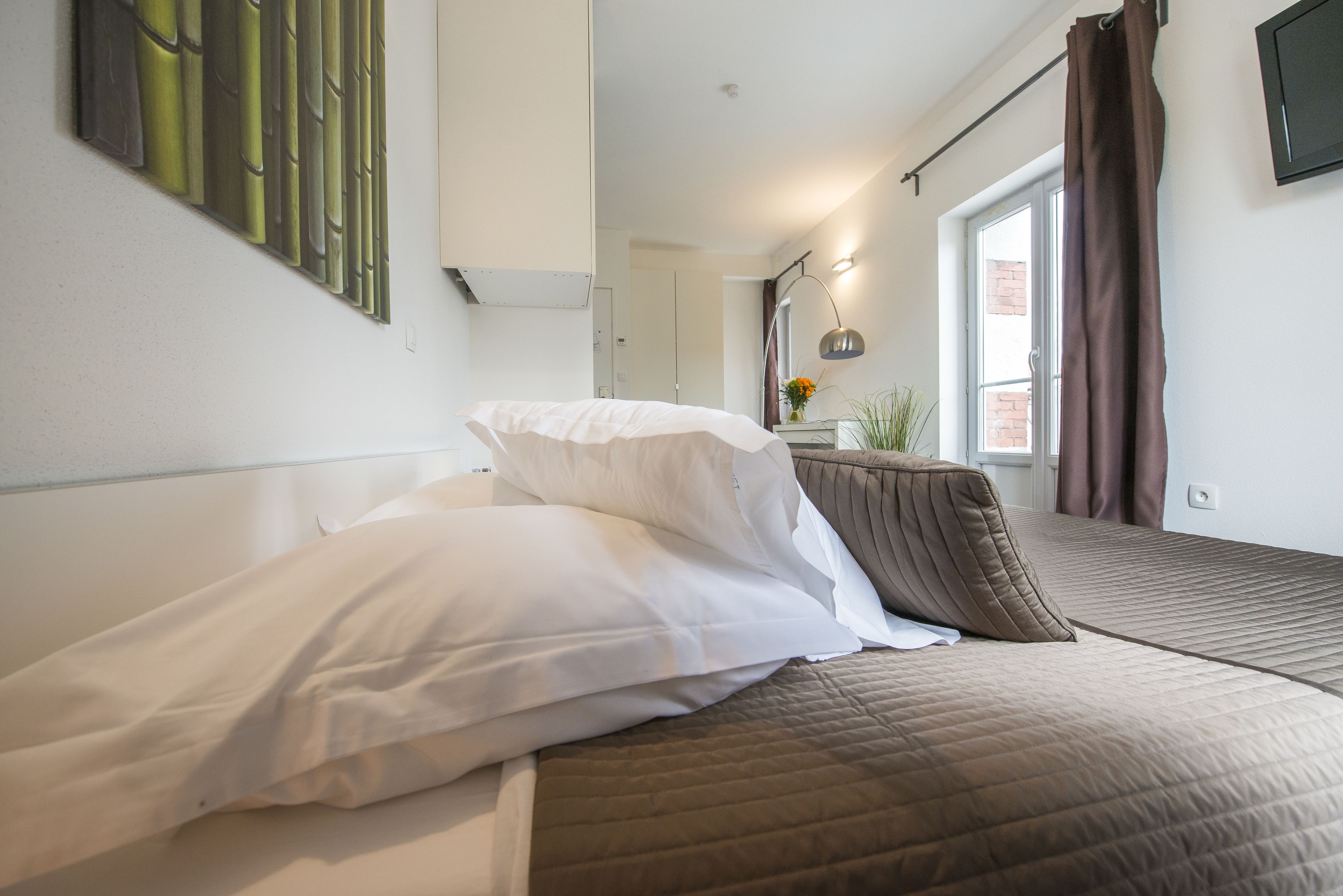 Ein Schlafzimmer mit Bett, einem Fenster mit Vorhängen, einem an der Wand befestigten Fernseher und einem Gemälde.