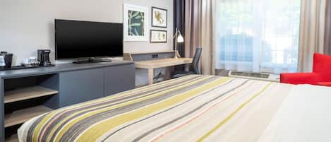 Hotellihuone, jossa on sänky, työpöytä tietokoneella, televisio ja punainen tuoli.