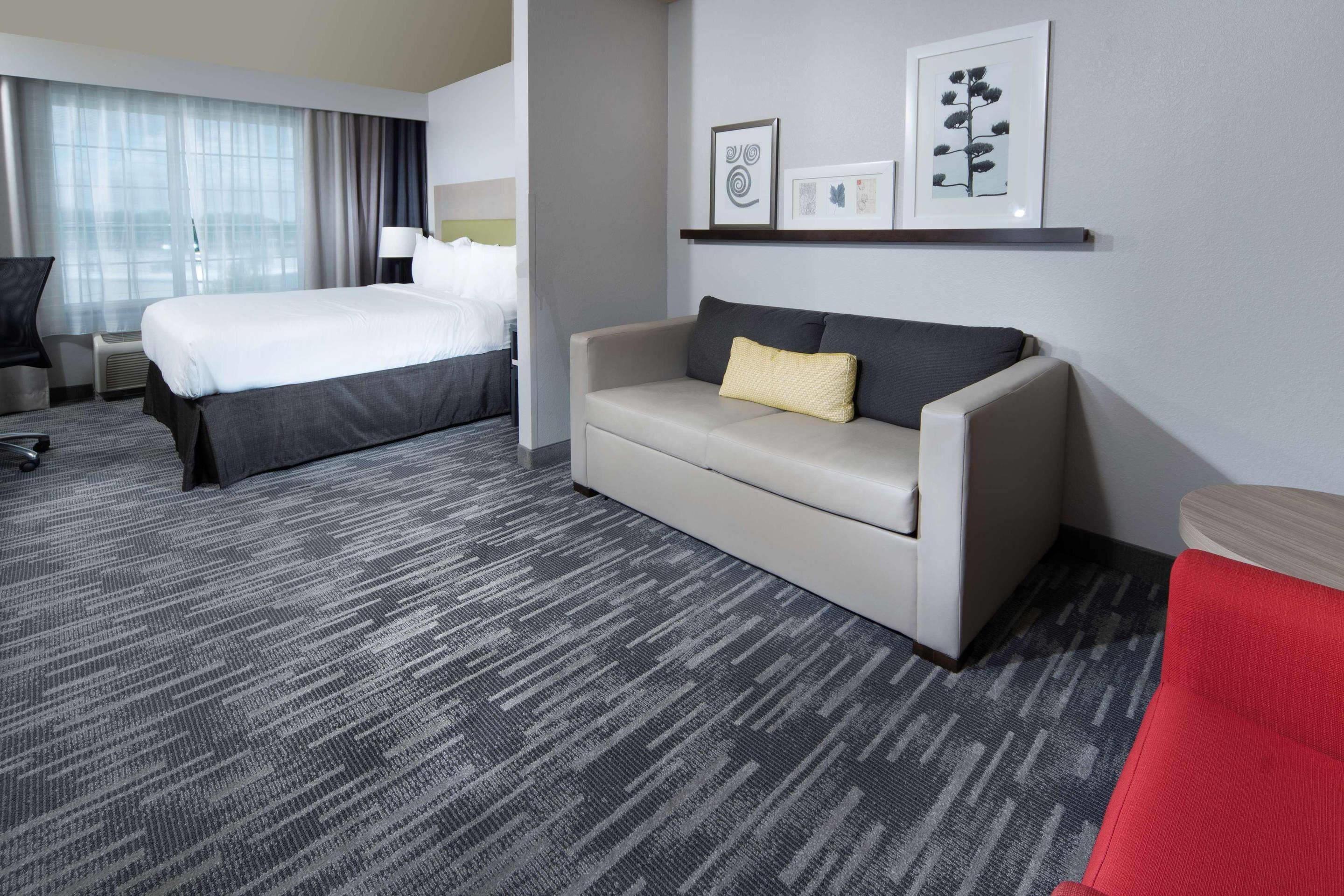 Habitación de hotel con una cama grande, un sofá, un escritorio y una silla. Se observan cuadros en la pared y una ventana con cortinas.