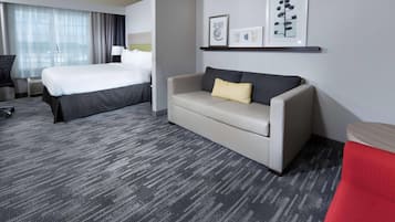 Ein Hotelzimmer mit einem großen Bett, einer Couch, einem Schreibtisch und einem Sessel. An der Wand befinden sich gerahmte Bilder und ein Fenster mit Vorhängen.