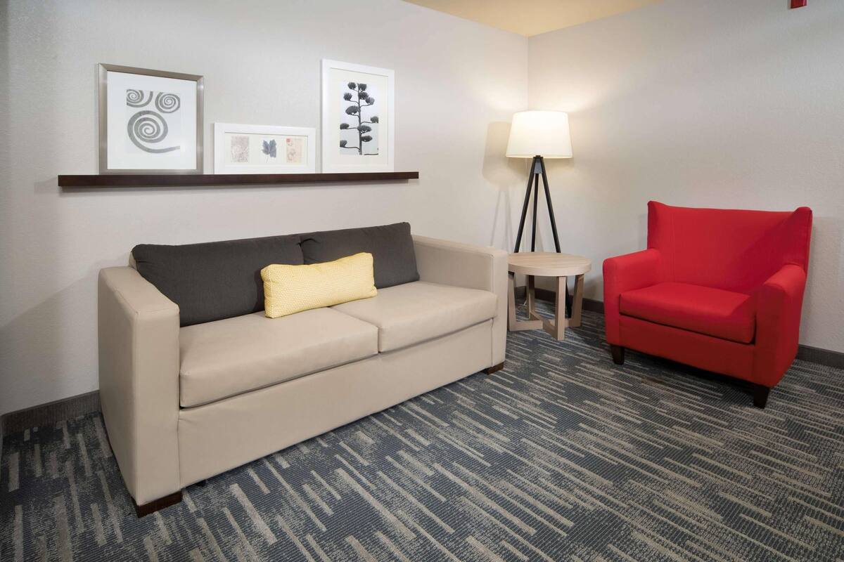 A hotel room with a beige sofa, a red armchair, a small table, a floor lamp, and artwork on the wall.