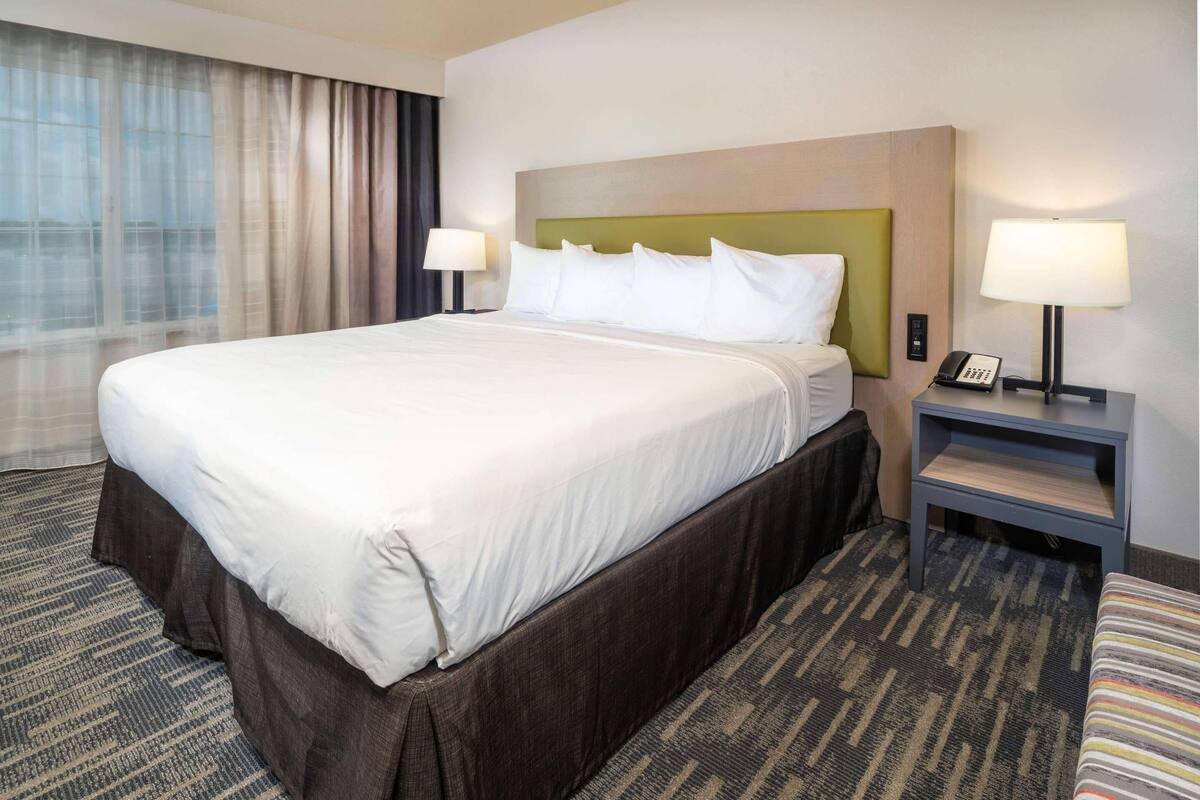 A hotel room with a large bed, two bedside tables with lamps, a carpeted floor, and a window with curtains.
