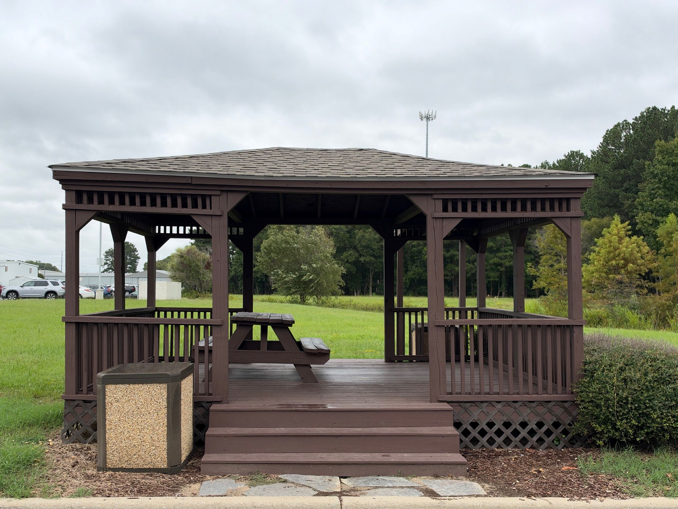 A wooden gazebo with a bench and steps leading up to it, surrounded by grass and trees.