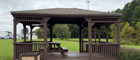 A wooden gazebo with a bench and steps leading up to it, surrounded by grass and trees.