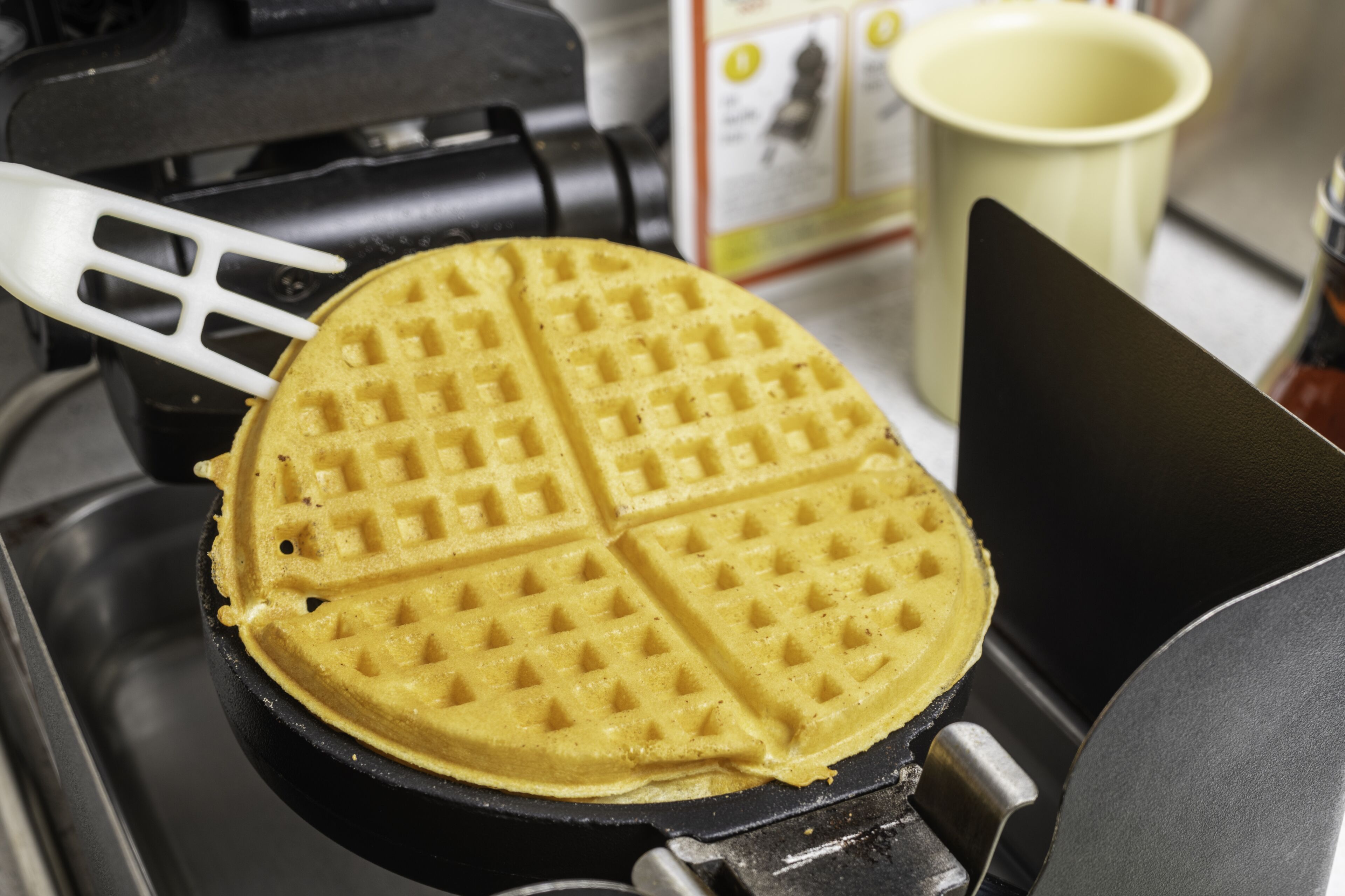 A waffle iron with a waffle inside, a spatula, and a cup in the background.
