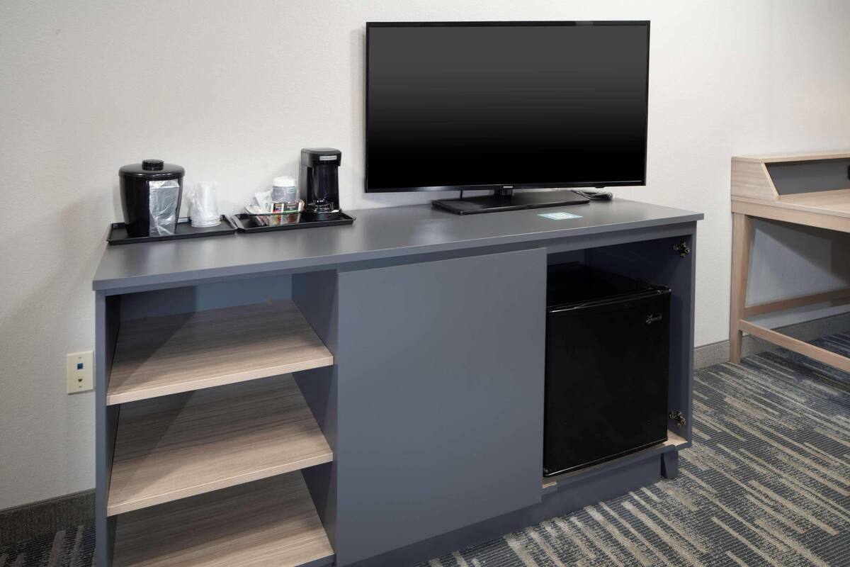 A modern hotel room with a grey console table, a flat-screen TV, a mini-fridge, and a coffee maker.