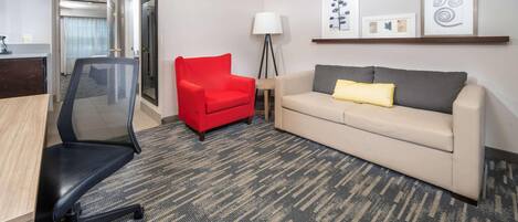 Una habitación de hotel con un escritorio, una silla roja, un sofá color beige con un cojín amarillo, una lámpara de pie y obras de arte enmarcadas en la pared.