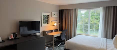 Une chambre d’hôtel avec un lit, un bureau et une chaise, une télévision et une fenêtre avec des rideaux.