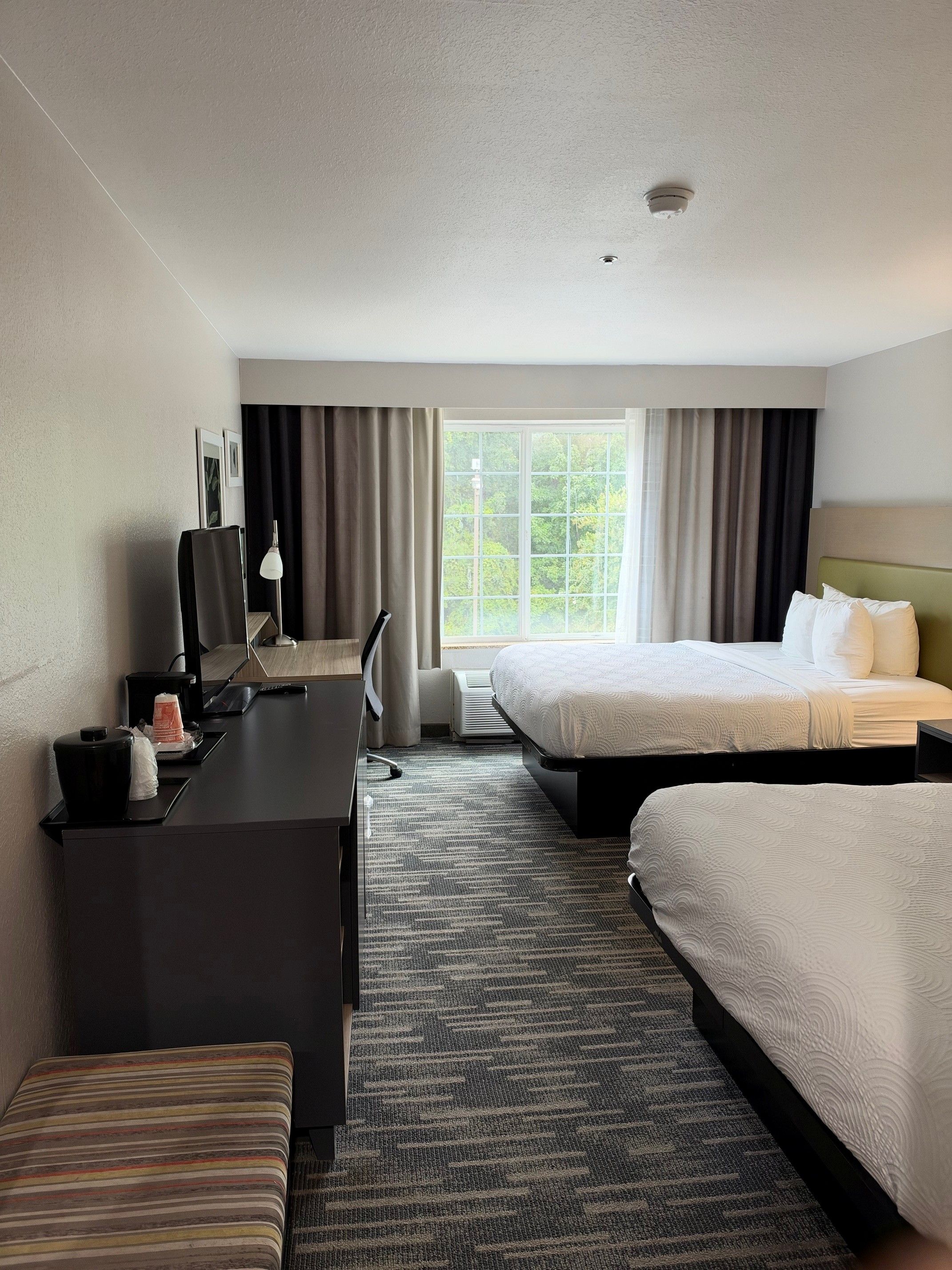 A hotel room with a desk, two beds, a window with curtains, and a ceiling light.