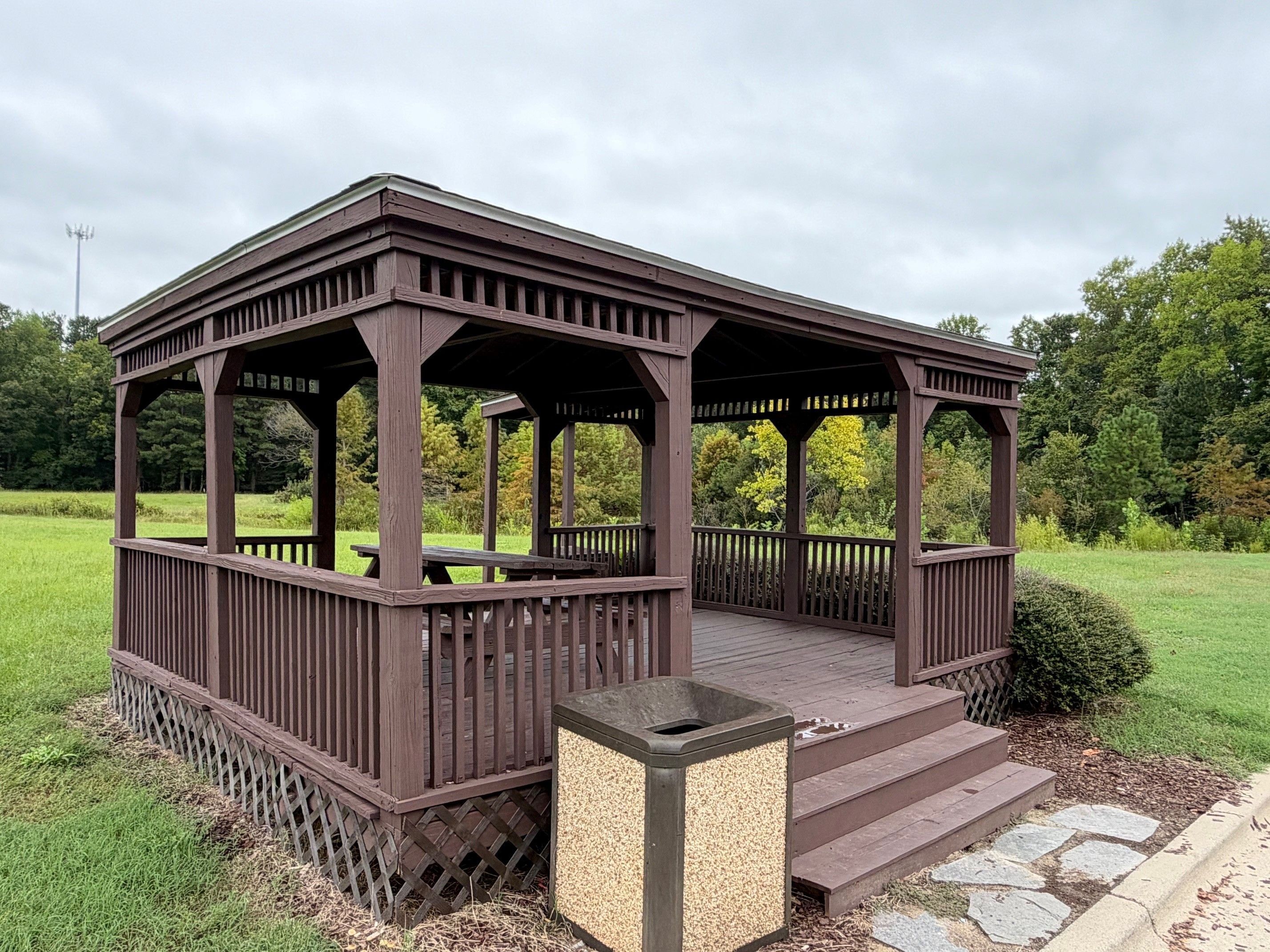 A wooden gazebo with a covered patio area, surrounded by a grassy field and trees.