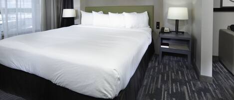 Ein Hotelzimmer mit einem großen Bett, zwei Nachttischen mit Lampen, einem Beistelltisch und Blick ins Freie durch ein Fenster mit durchsichtigen Vorhängen.