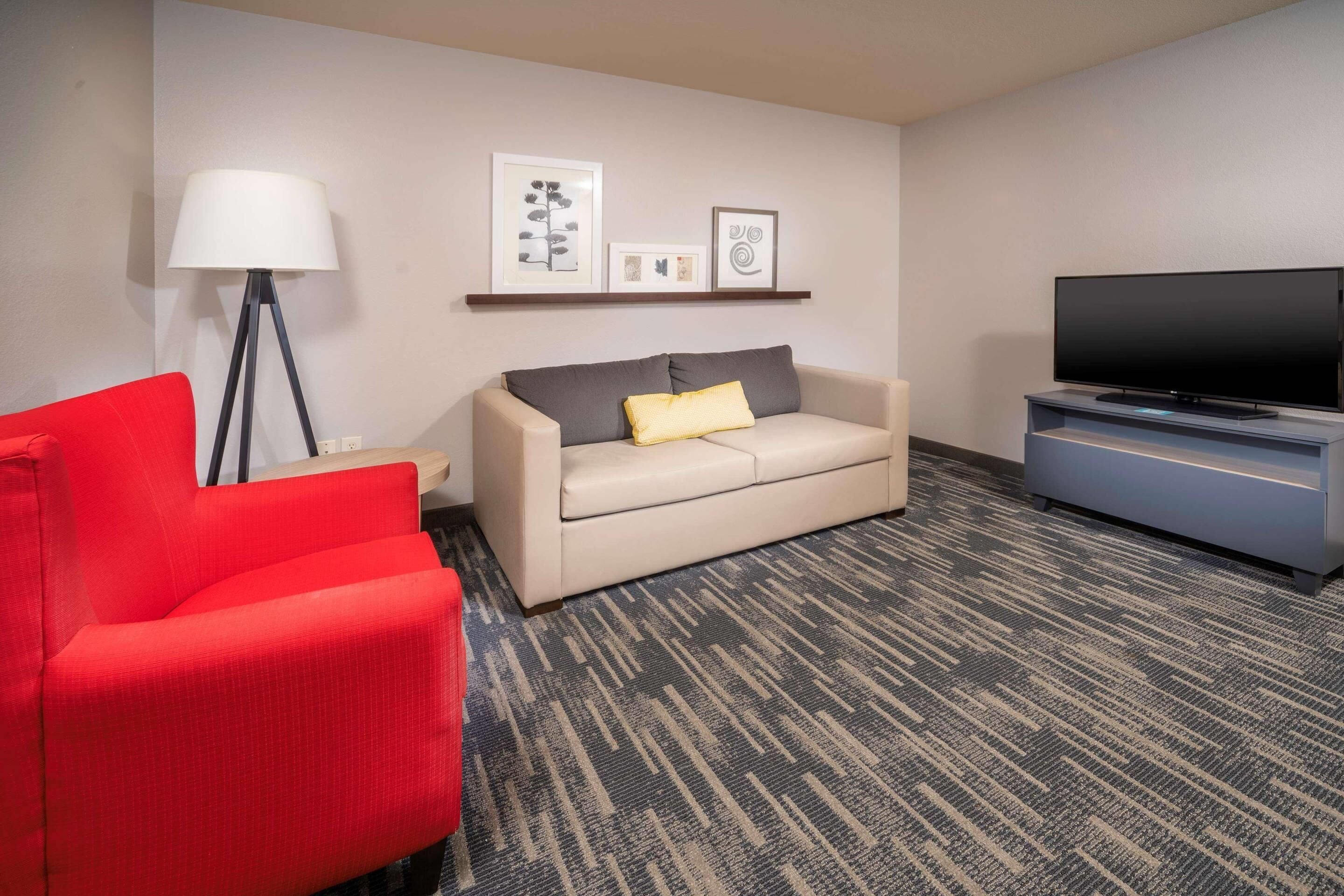 A hotel room with a red armchair, a beige sofa with a yellow pillow, a floor lamp, a flat-screen TV, and framed artwork on the wall.