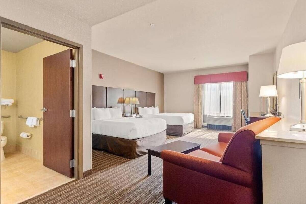 Une chambre d’hôtel avec un lit, deux tables de chevet, un bureau, une chaise et une salle de bain visible à travers une porte ouverte.