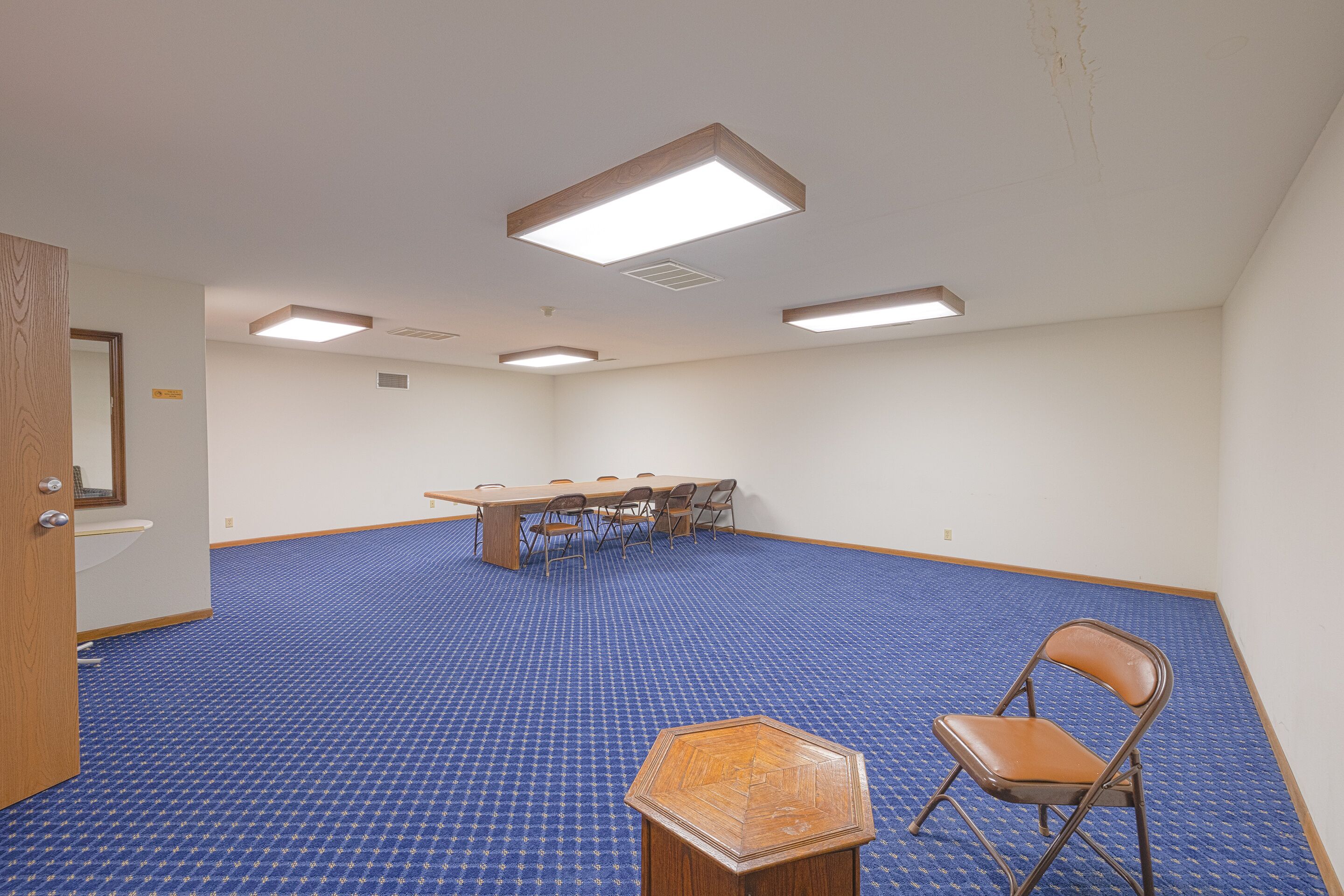 Uma sala de conferências com carpete azul, uma mesa de madeira com cadeiras e um abajur hexagonal.