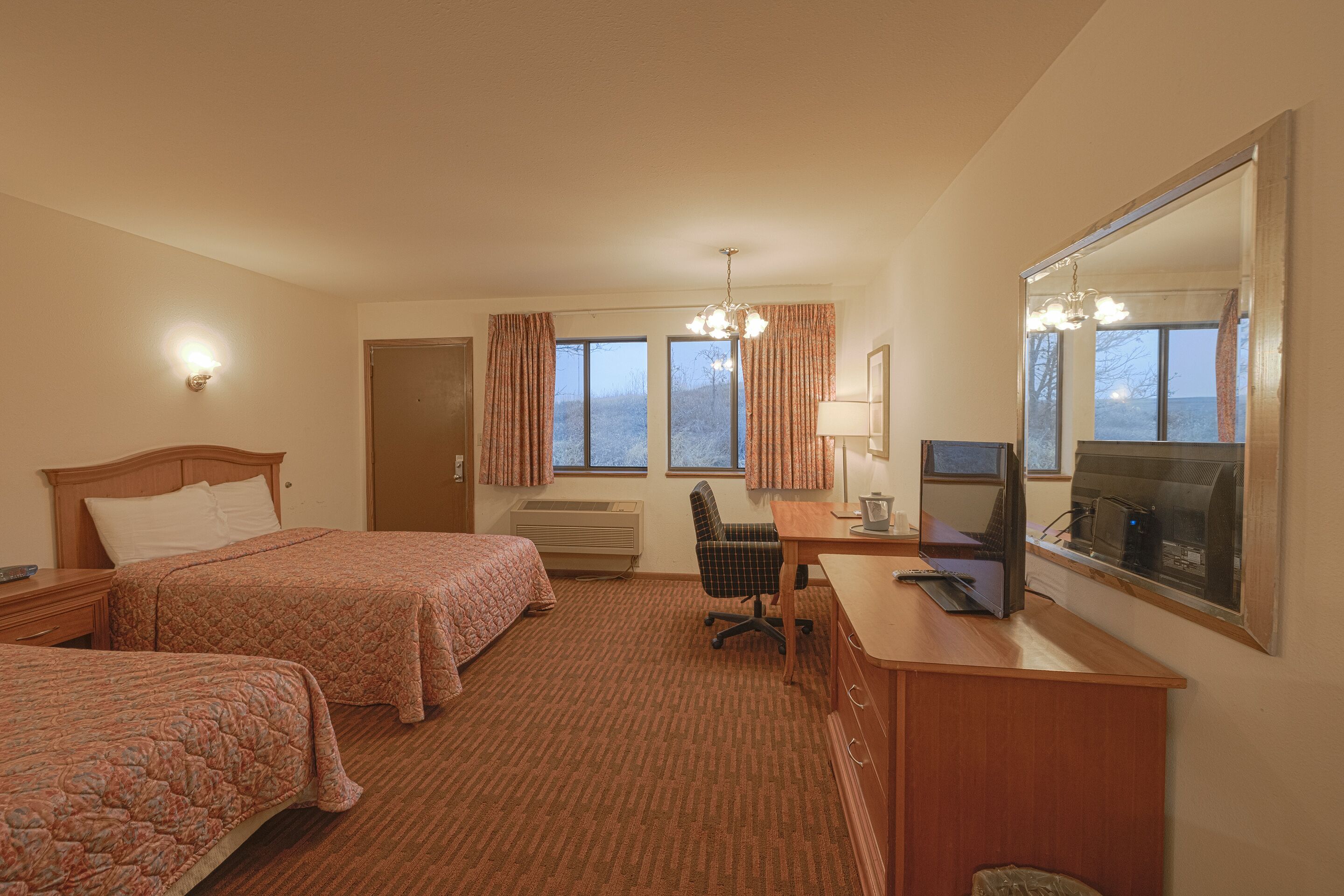 Habitación de hotel con dos camas, un escritorio con ordenador, una silla, un espejo grande y vistas al exterior a trav és de las ventanas.