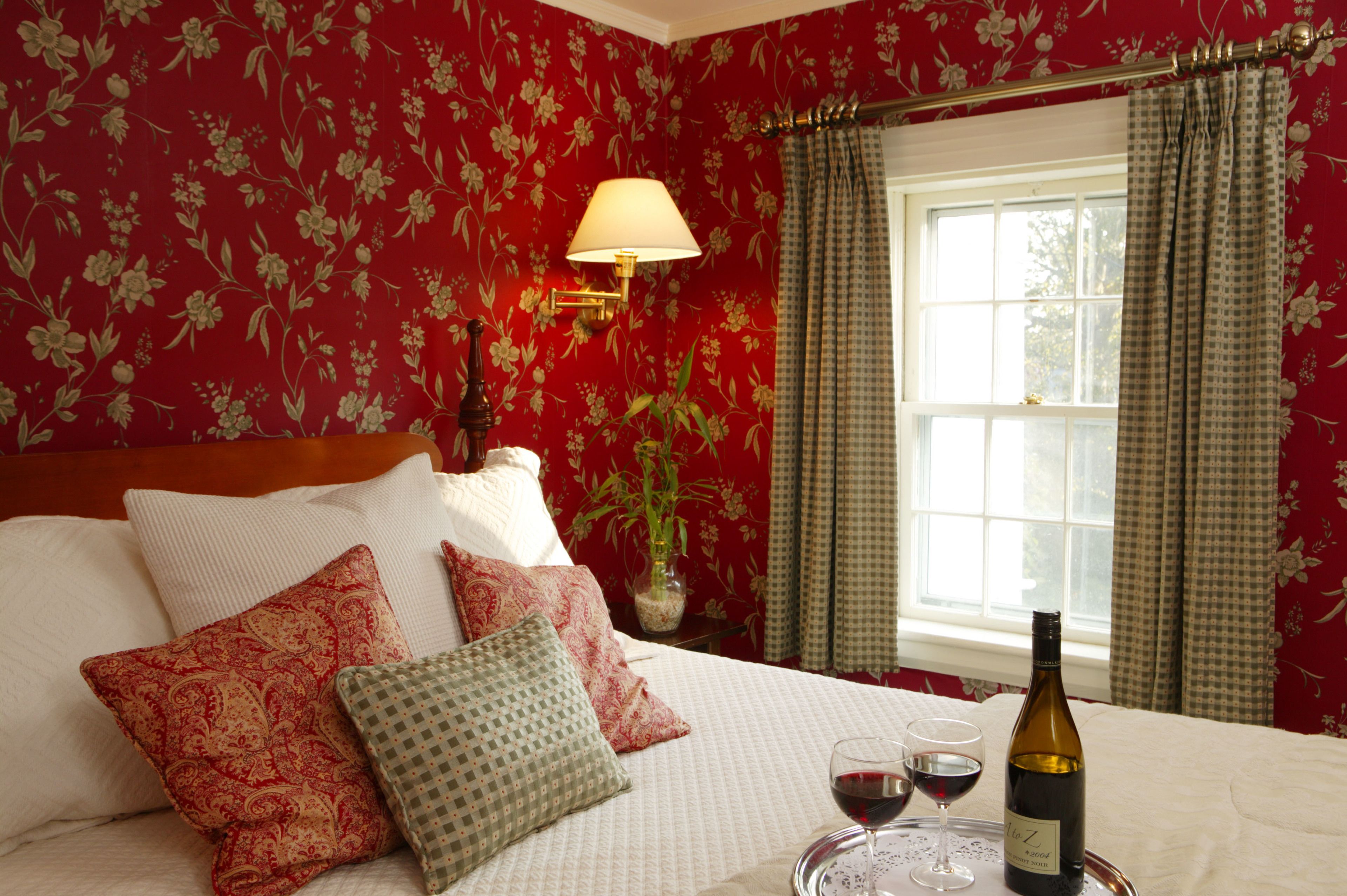 A hotel room with floral wallpaper, a bed with multiple pillows, a bedside table with a lamp, a window with curtains, and a tray with wine glasses and a bottle.