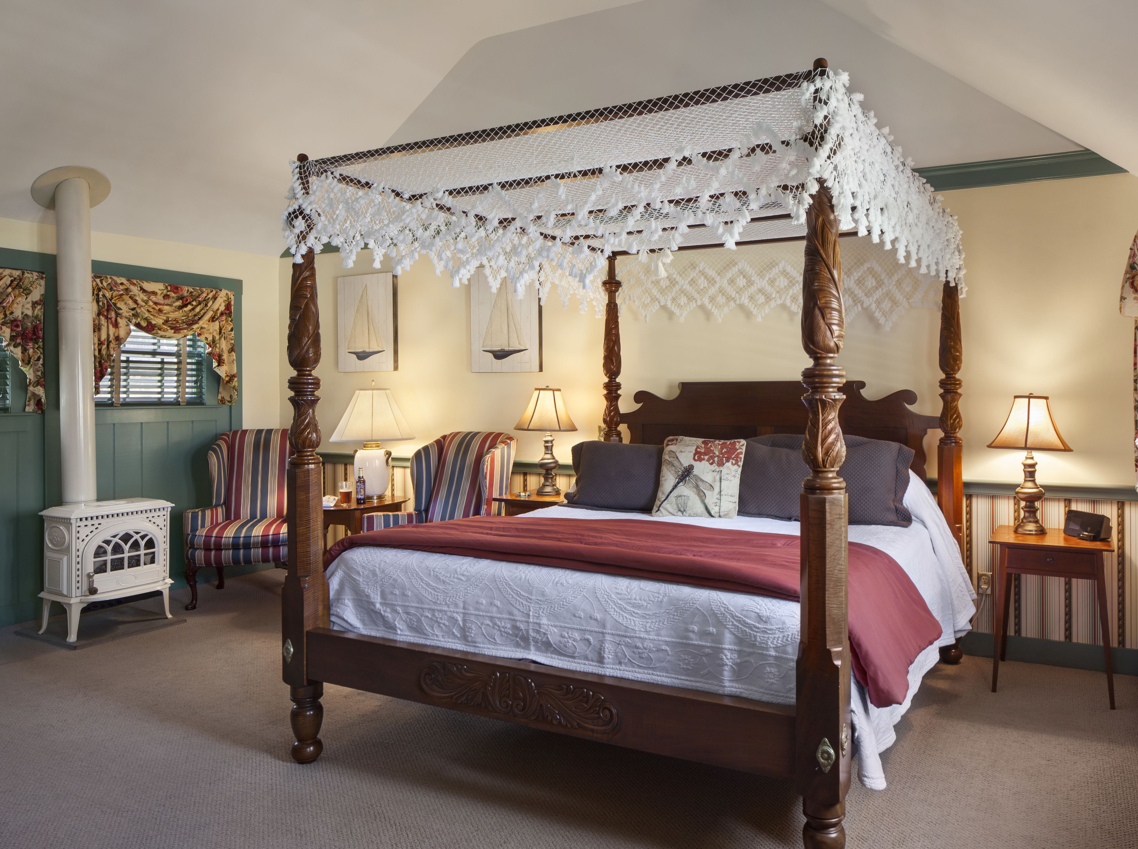 A four-poster canopy bed with a lace canopy, a wooden headboard, and a red bedspread. There are two bedside tables with lamps, a striped armchair, and a small white cabinet.