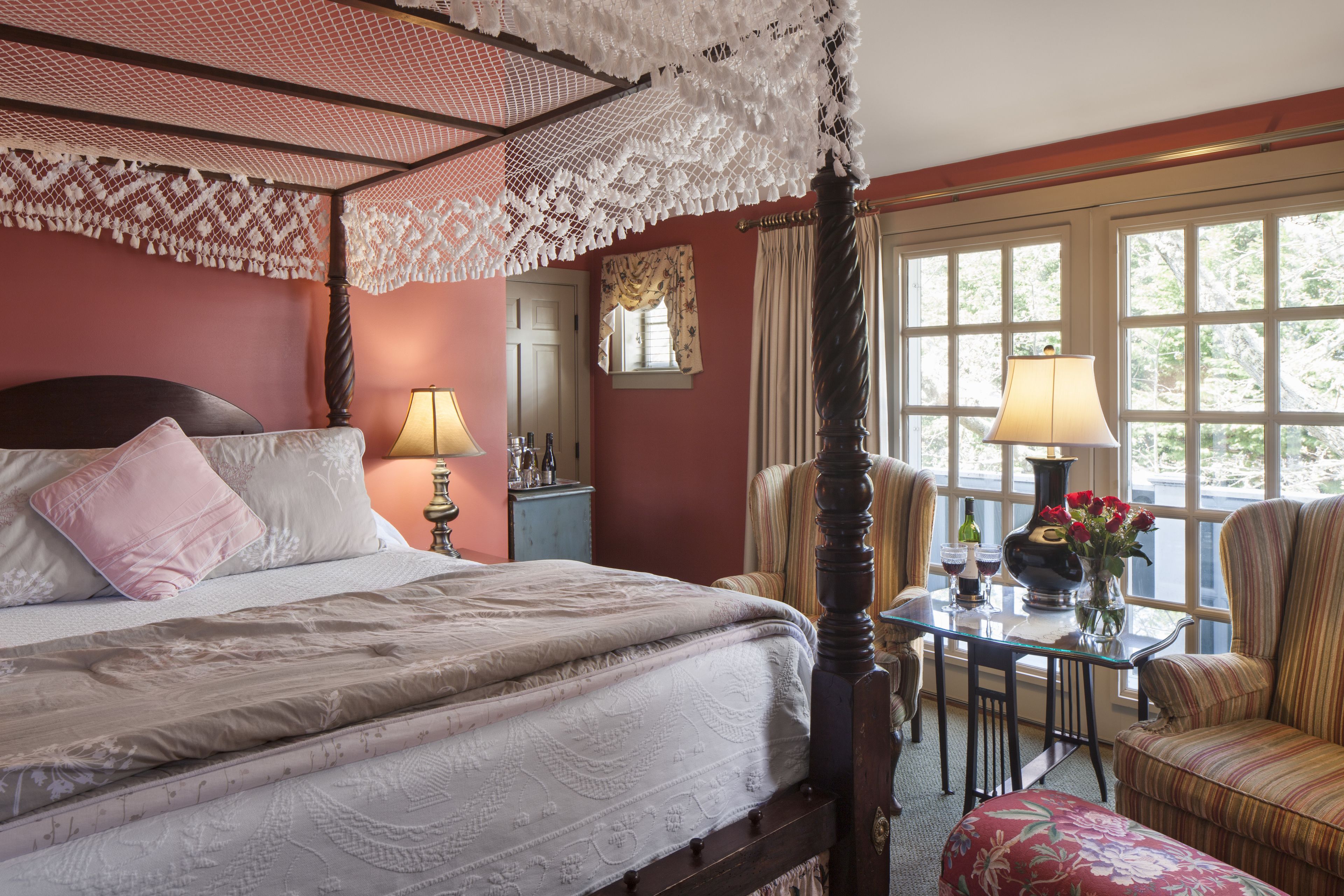 A bedroom with a four-poster bed, a small table with a lamp, a chair, and a view of the outdoors through large windows.