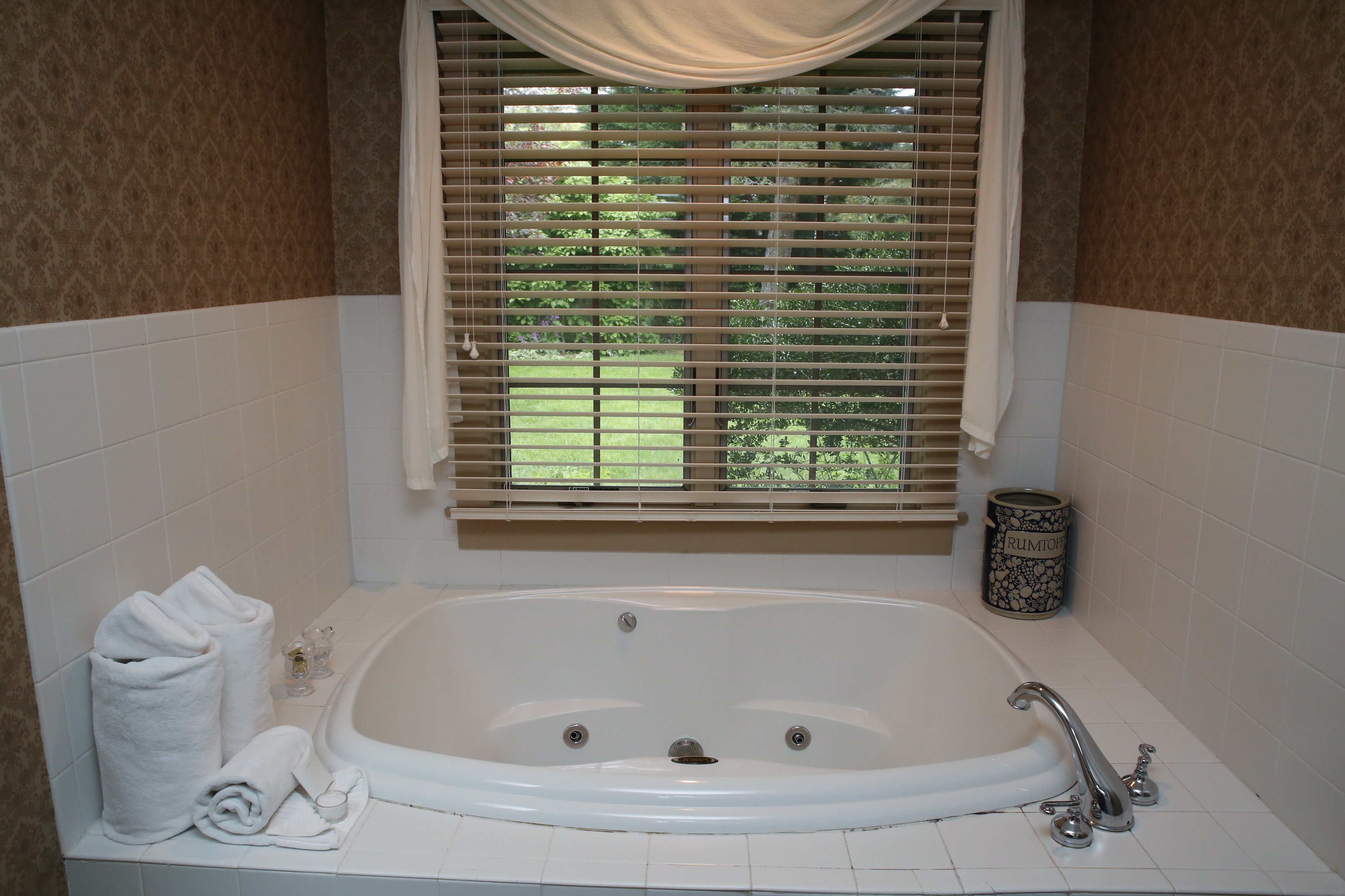 A bathroom with a large bathtub, white tiled walls, a window with blinds, and a trash can.