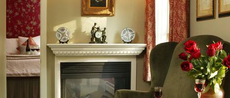 Una sala de estar acogedora con chimenea, un sillón verde, una mesita con libros y un jarrón de rosas rojas, y una colcha floral roja al fondo.