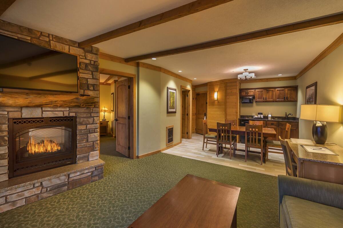 A cozy living room with a stone fireplace, wooden beams, a dining area, and a kitchen with wooden cabinets.