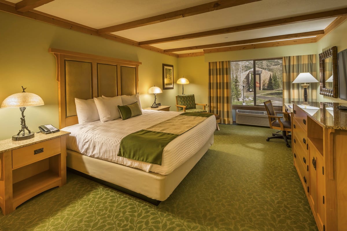A hotel room with a large bed, a desk, a chair, and a dresser. There is a window with curtains and a view of the outside.